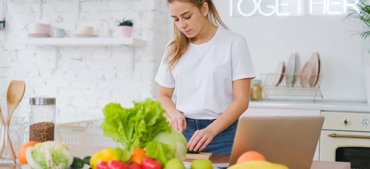 Woman in white t-shirt preparing fresh vegetables in a bright kitchen with a laptop.